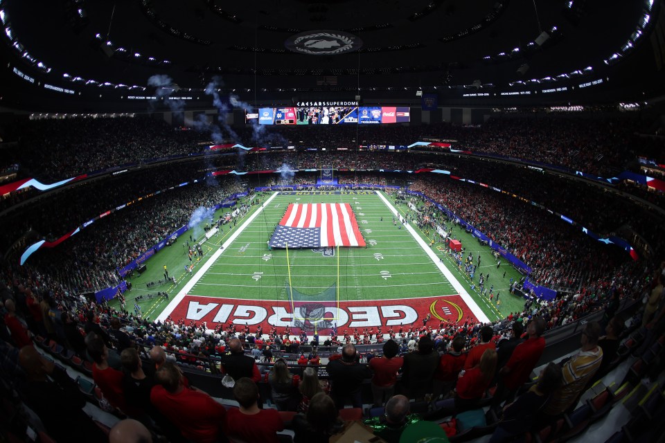 A wide shot of a football stadium during a pre-game ceremony, showing a large American flag on the field and spectators in the stands.